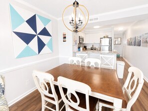 A dining area with a wooden table and white chairs, a chandelier, and a kitchen in the background. - Dunescape Villas 108 (Atlantic Beach)