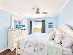 A bedroom with a bed, a dresser, a ceiling fan, a television, and a view of the beach. - Dunescape Villas 108 (Atlantic Beach)