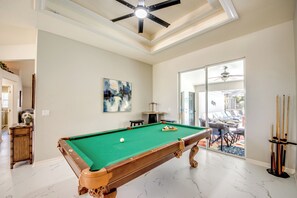 Uma mesa de bilhar em um ambiente com ventilador de teto, um quadro na parede e vista através de uma porta de vidro.