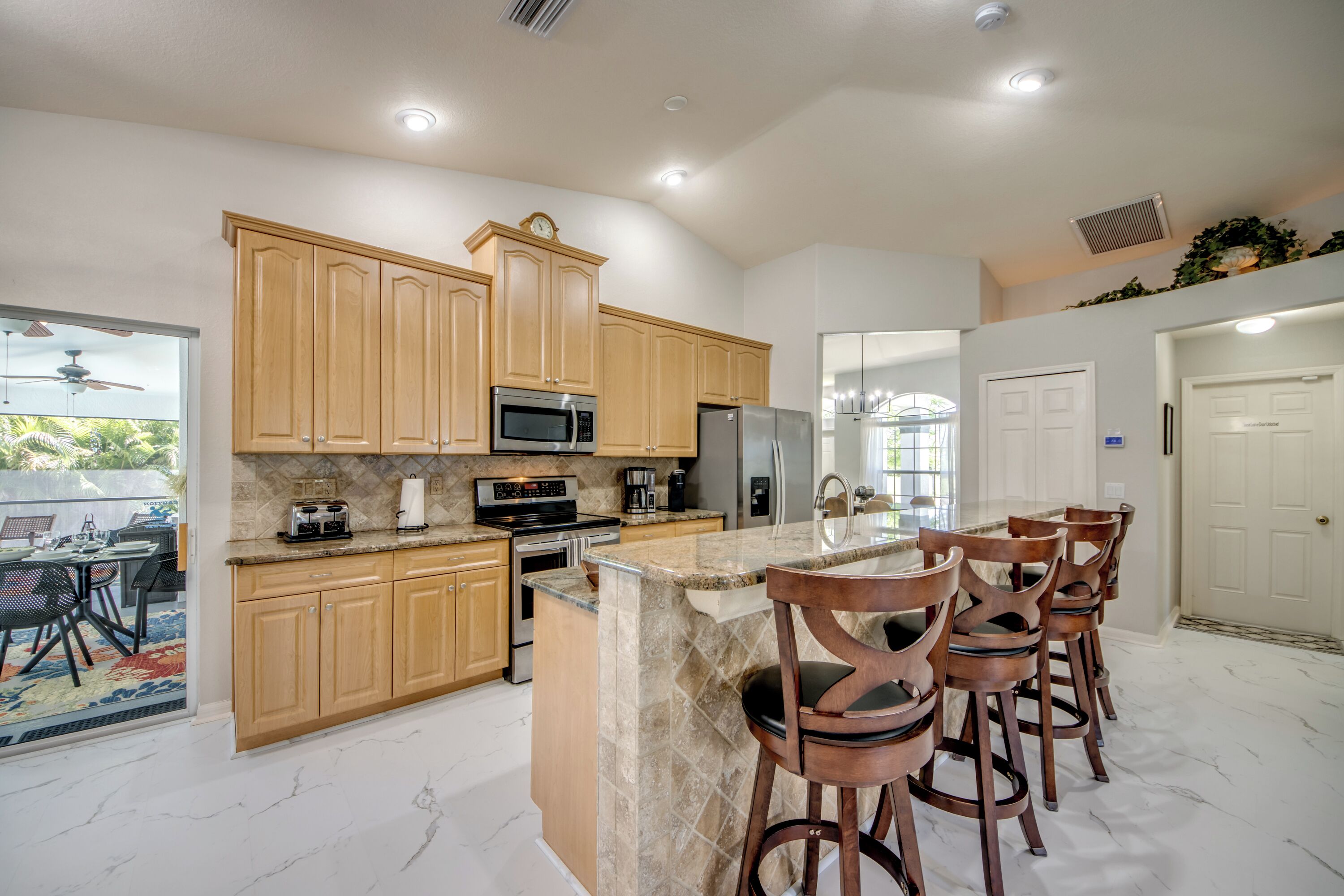 A modern kitchen with wooden cabinets, a central island with bar stools, stainless steel appliances, and a view to an outdoor patio.