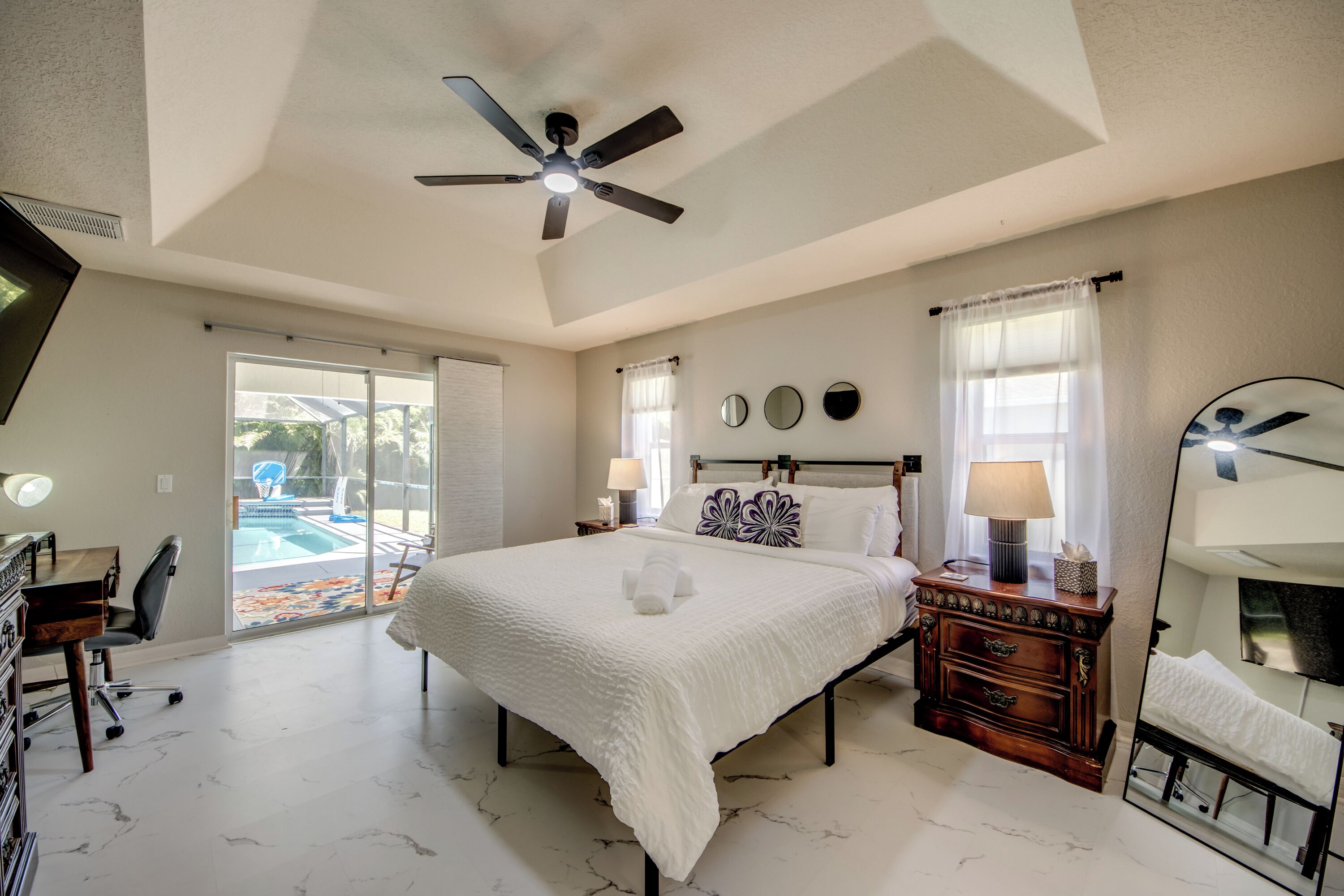 A bedroom with a bed, bedside tables, a mirror, a ceiling fan, and a view of a swimming pool.
