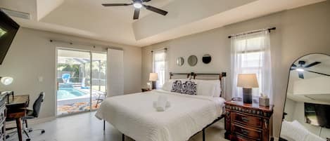 A bedroom with a bed, bedside tables, a mirror, a ceiling fan, and a view of a swimming pool.
