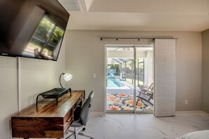 Ein Zimmer mit Schreibtisch aus Holz, einem Stuhl, einem an der Wand befestigten Fernseher, einem Deckenventilator und einer Glastür zum Poolbereich.