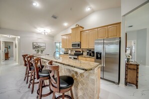 A modern kitchen with wooden cabinets, a stainless steel refrigerator, a marble countertop, and bar stools. - Tropical Paradise-Salt Water Pool-Billiards-Pool Volley/Basket Ball-Sleeps 8 (Cape Coral)