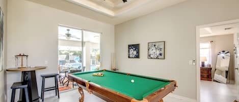 A pool table with a ceiling fan, a billiards rack, and a bar area with stools.