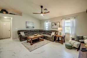 Ein Wohnzimmer mit einer Eckcouch, einem Couchtisch, einem Beistelltisch mit Lampe, einem Sessel und einem Deckenventilator.