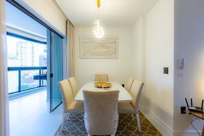 Un espace repas moderne avec une table blanche, des chaises rembourrées, une œuvre d’art encadrée et une vue à travers une porte vitrée.