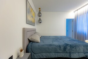 A bedroom with a bed, a bedside table, a lamp, a painting, and a door leading to another room. - Apartment w/ Gourmet Balcony | 180m from Perequê Beach (Porto Belo)