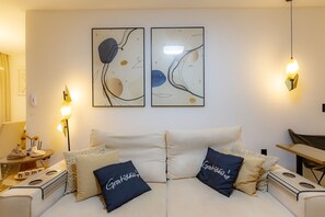 A modern living room with a sofa, pillows, a small table with a lamp, and abstract wall art. - Apartment w/ Gourmet Balcony | 180m from Perequê Beach (Porto Belo)