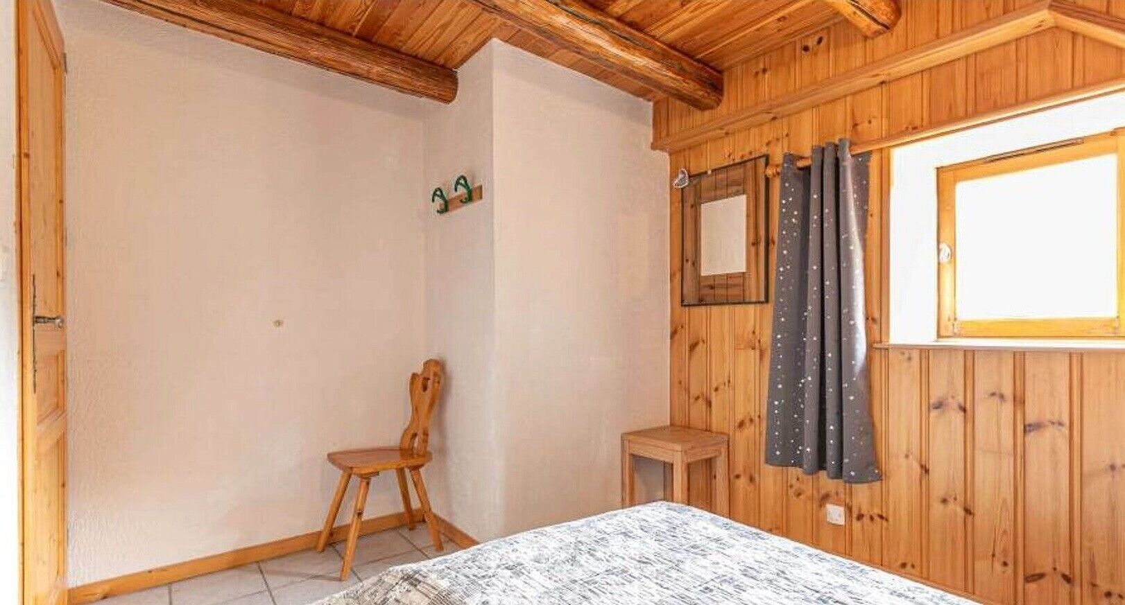 Une pièce aux murs en bois, avec un lit, une chaise, une petite table et une fenêtre avec des rideaux.