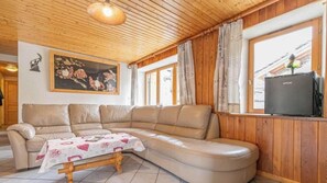 Una sala de estar acogedora con un amplio sofá en forma de L, una mesita con mantel, paredes de madera, un ventilador de techo y un gran ventanal con cortinas.
