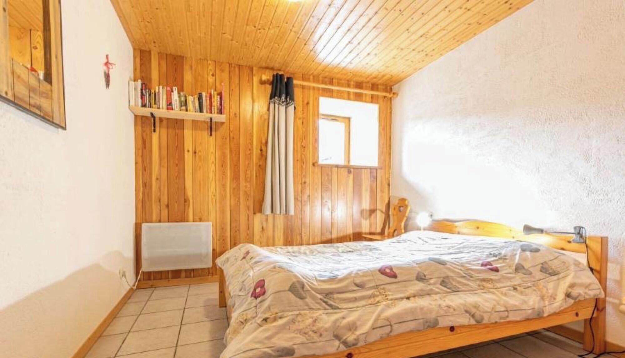 A room with a wooden ceiling, a bed with a patterned bedspread, a window, and a shelf with books.