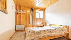 A room with a wooden ceiling, a bed with a patterned bedspread, a window, and a shelf with books.