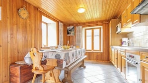 A wooden interior with a dining table, chairs, a bench, a clock, and a kitchen area with cabinets and appliances.