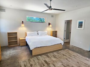 Un dormitorio con cama, mesitas de noche, ventilador de techo, un cuadro y una puerta.