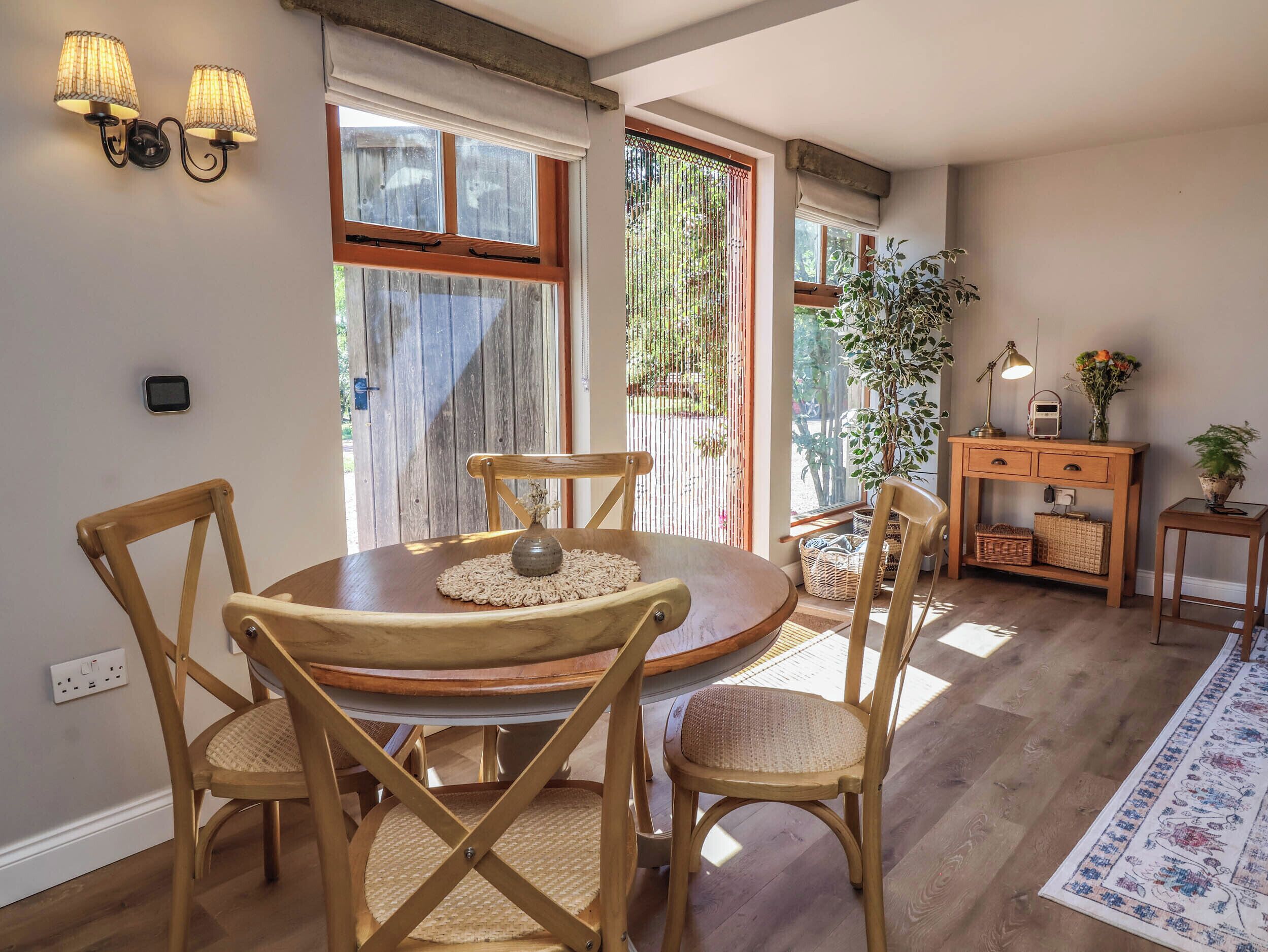 A dining area with a round wooden table, four wooden chairs, a small vase, and a wall-mounted light fixture.