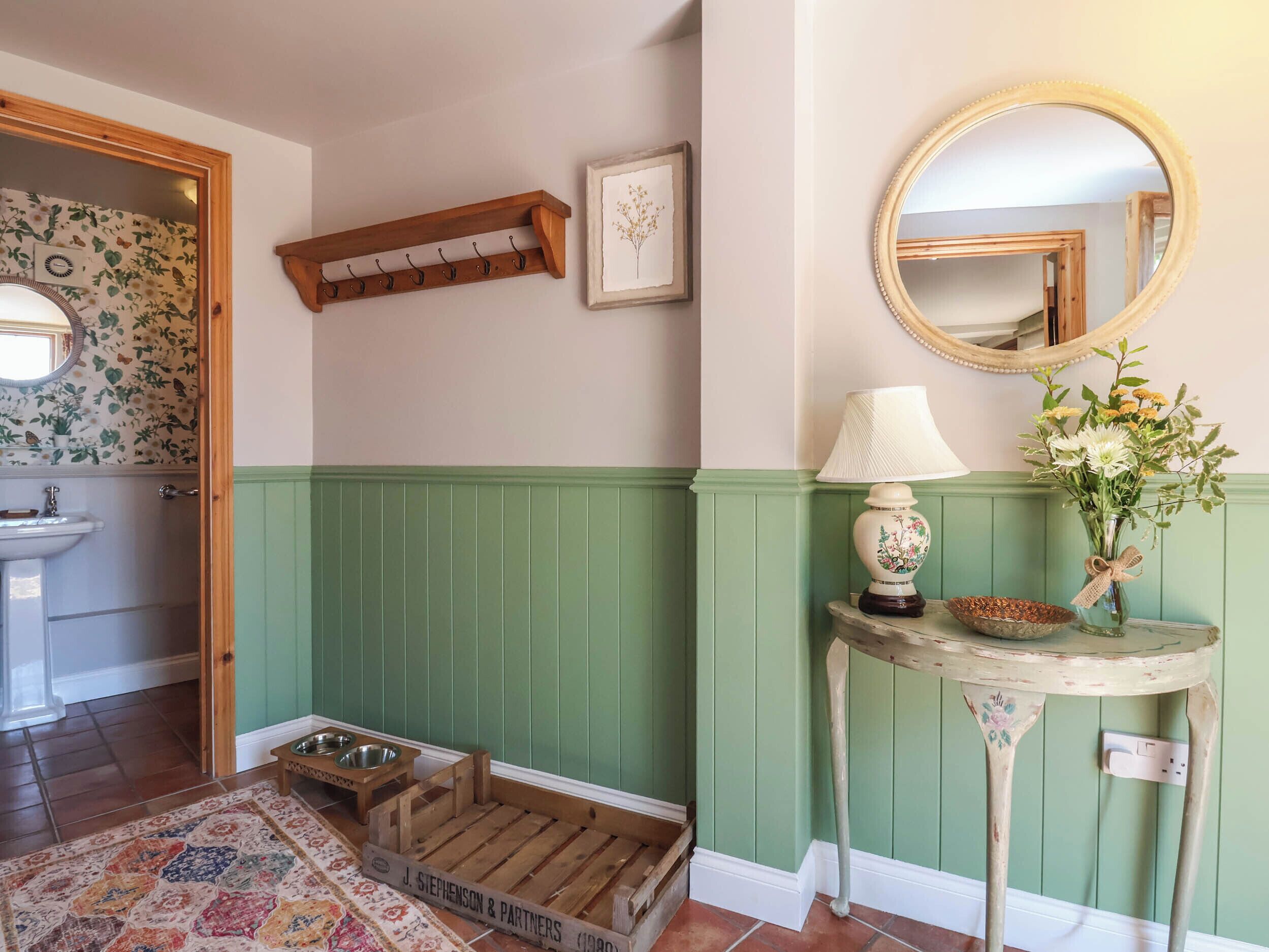 A room with a wooden coat rack, a framed picture, a round mirror, a table with a lamp, a vase with flowers, a pet feeding station, and a patterned rug.