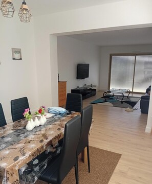 Ein Essbereich mit Tisch für vier Personen, einer Couch, einem Fernseher und einem Fenster mit Jalousien.