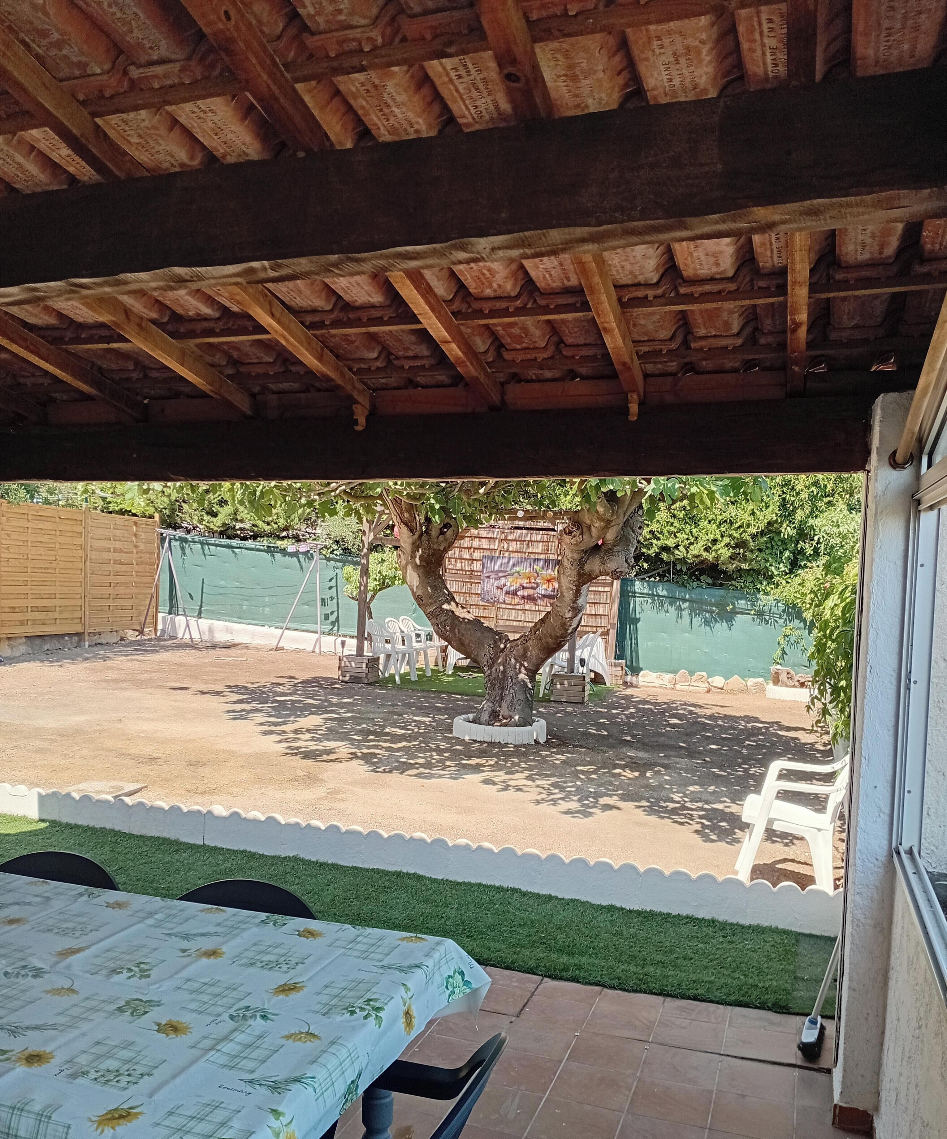 Ein überdachter Patio-Bereich mit Tisch und Stühlen, ein Baum und ein grüner Zaun im Hintergrund.