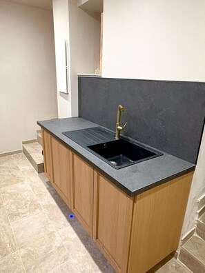 A kitchen with a black countertop, a gold faucet, and wooden cabinets. - The Little House — An Air-Conditioned Provençal Retreat (Caumont-sur-Durance)