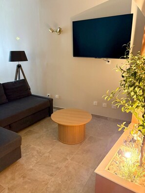A living room with a dark-colored sofa, a round wooden coffee table, a floor lamp, a wall-mounted TV, and a potted plant. - The Little House — An Air-Conditioned Provençal Retreat (Caumont-sur-Durance)