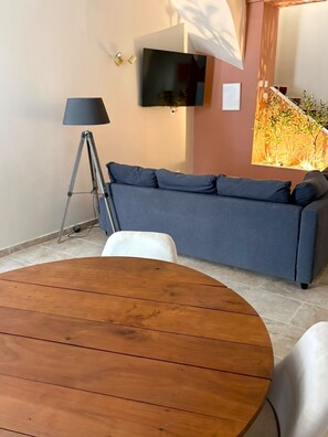 A room with a round wooden table, a blue sofa, a floor lamp, and a flat-screen TV mounted on the wall. - The Little House  — An Air-Conditioned Provençal Retreat (Caumont-sur-Durance)