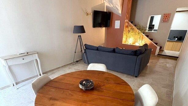 Un comedor con una mesa redonda de madera, sillas blancas tapizadas, un pequeño cuenco decorativo y un sofá moderno al fondo.