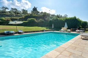 Una piscina rectangular d'aigua transparent i blava, envoltada per un solàrium de pedra. Hi ha gandules i ombrel·les al costat de la piscina.