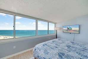A bedroom with a large window overlooking a beach, a bed with a patterned bedspread, and a wall-mounted TV displaying a vacation advertisement. - Antigua 1103 by Seaside Vacations & Sales (Ocean City)