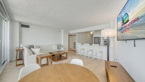 Uma sala de estar moderna com sofá, mesa de centro e área de jantar. Cozinha visível ao fundo, com eletrodomésticos brancos e uma grande janela.