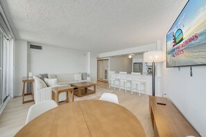 A modern living room with a sofa, coffee table, and dining area. There is a kitchen in the background with white appliances and a large window. - Antigua 1103 by Seaside Vacations & Sales (Ocean City)