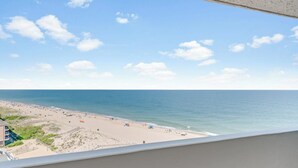 Vista da praia a partir de uma varanda, com céu claro, praia de areia e oceano.