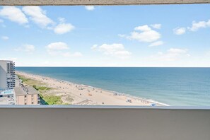 Blick auf einen Strand vom Balkon aus, mit blauem Himmel, Sandstrand und einem Gebäude im Vordergrund.
