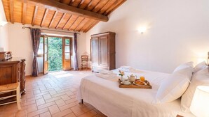 A bedroom with a bed, a wooden wardrobe, a chair, a table with breakfast items, and a view of greenery outside.