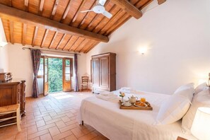 Un dormitorio con cama, armario de madera, silla, mesa con provisiones para el desayuno y vista a zonas verdes.