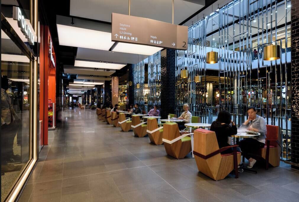 A modern indoor dining area with tables and chairs, a directional sign overhead, and a glass wall on one side.