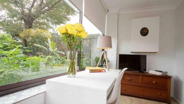 Een moderne kamer met een wit bureau, een vaas met gele bloemen, een lamp, een televisie en een boekenkast.