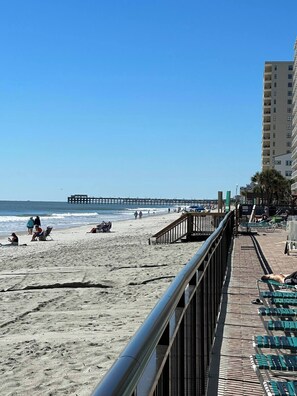 A beach with a pier in the distance, a walkway with chairs, and a building with balconies. - Turning Tides: 1 bed, 2 bath direct luxury oceanfront condo in Garden City (Murrells Inlet)