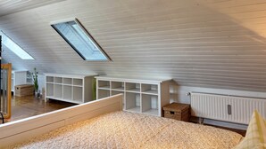 Una habitación con techo inclinado, tragaluces, estanterías empotradas blancas, un radiador y una cama con cubierta texturizada.