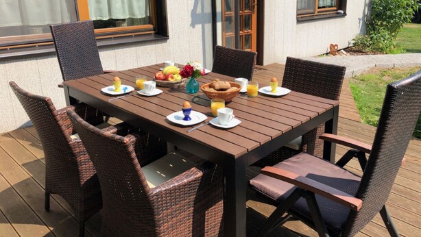 Una terraza de madera con una mesa lista para el desayuno, sillas de mimbre y un pequeño jardín.