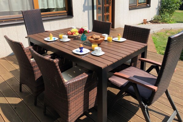 A wooden deck with a table set for breakfast, wicker chairs, and a small garden area.