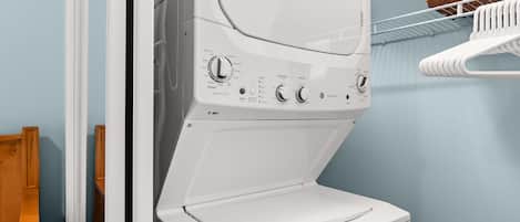 A front-loading white washer and dryer unit with control knobs, a wooden bench, and a metal shelf with a cushion.
