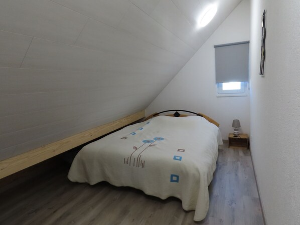 Ein kleiner, schräger Raum mit einem Einzelbett, einem Nachttisch und einem Fenster.