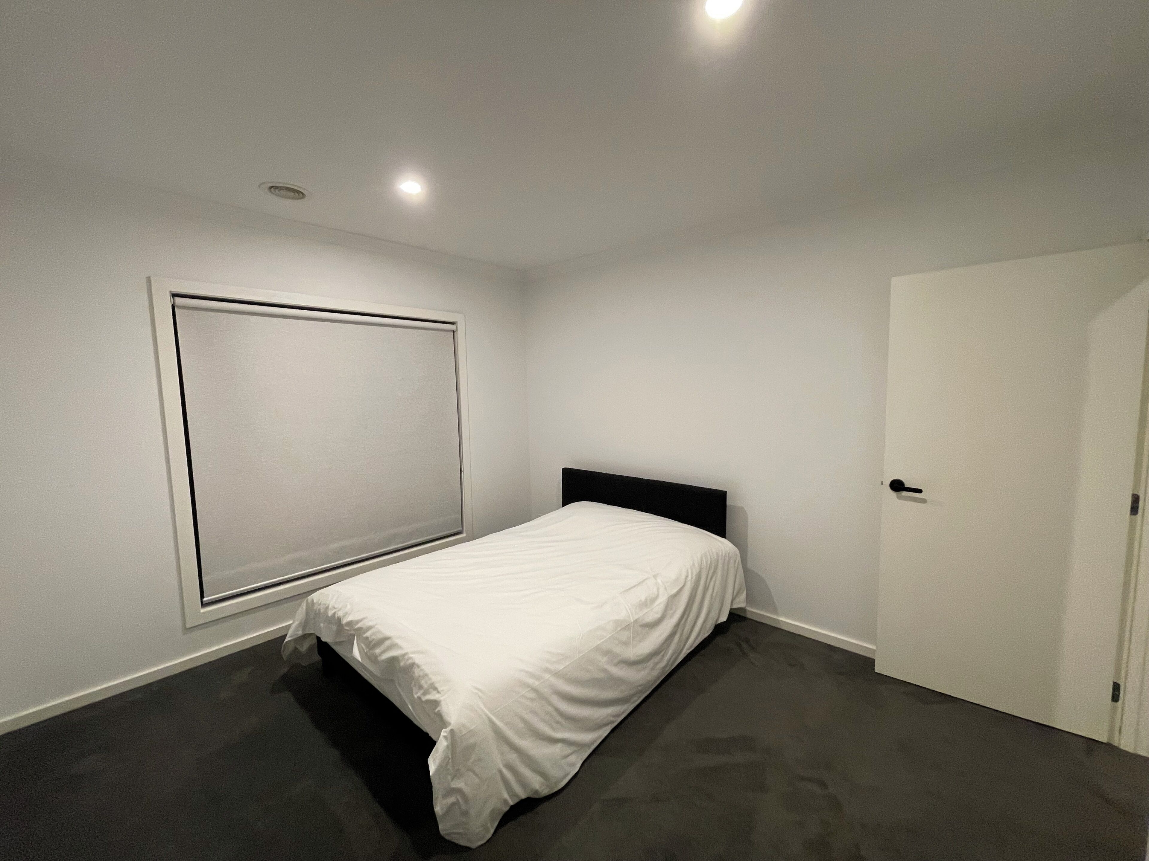 A bedroom with a single bed, a window with blinds, a door, and a carpeted floor.
