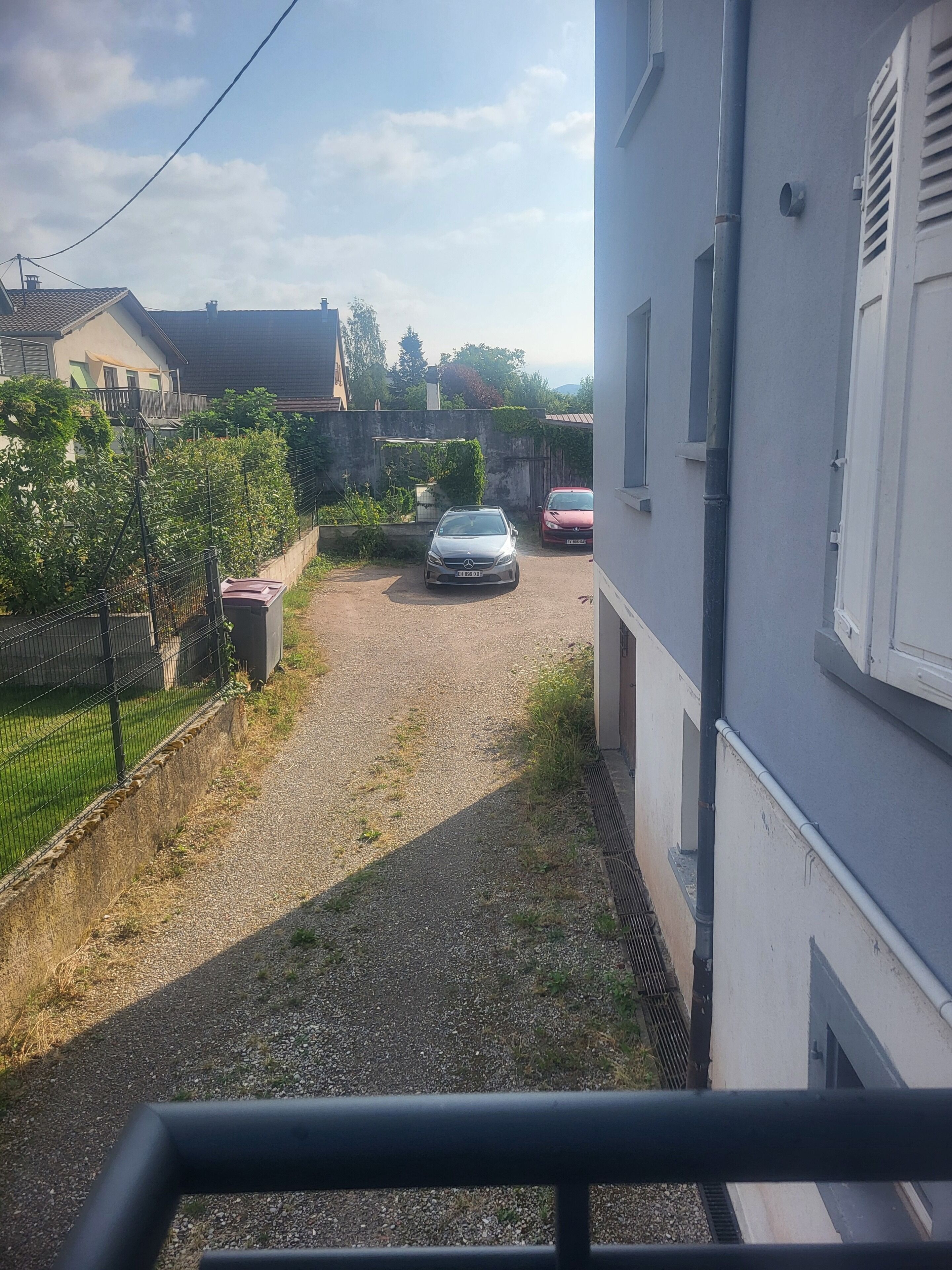Une rue résidentielle avec deux voitures garées, une allée gravillonnée et une clôture.