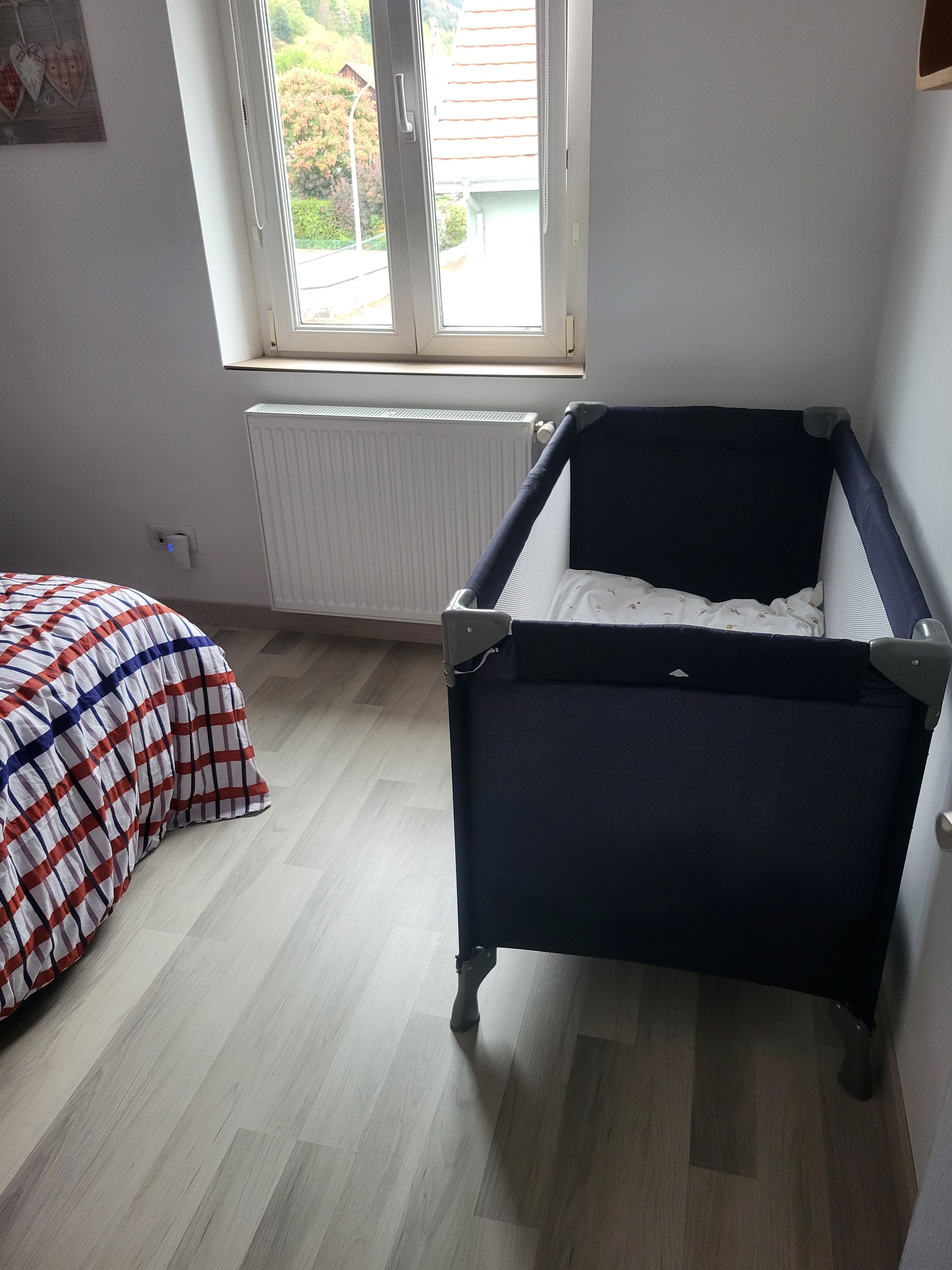 Une chambre avec un berceau, un radiateur, une fenêtre et un lit recouvert d’une couverture à carreaux.