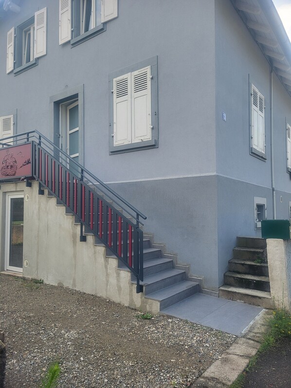 Un edificio con fachada gris, una escalera de hormigón con barandillas rojas y contraventanas blancas en las ventanas.