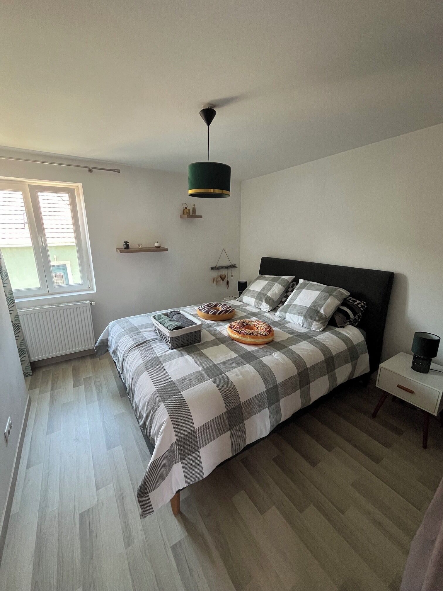 A bedroom with a bed, a bedside table, a hanging light fixture, a window, and a radiator.