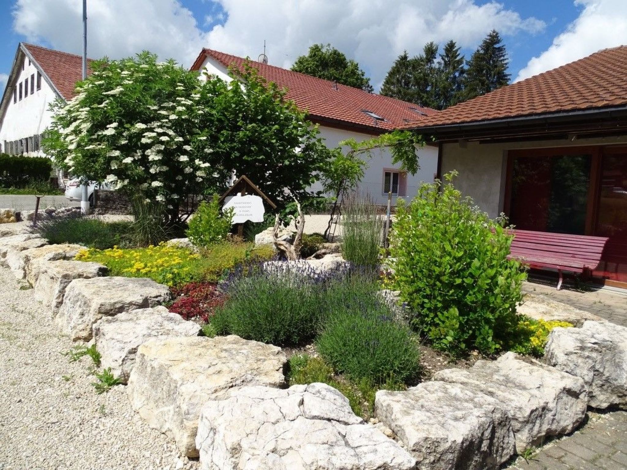 Um edifício com telhado de telhas vermelhas, rodeado por um jardim com diversas plantas e uma parede de pedra.