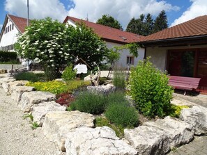 Ein Gebäude mit roten Ziegeln auf dem Dach, umgeben von einem Garten mit verschiedenen Pflanzen und einer Steinmauer.